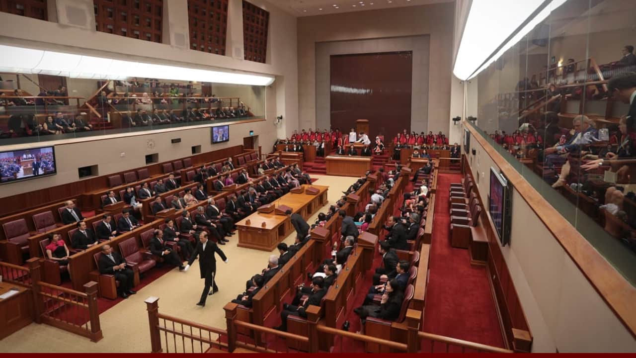 A large parliamentary chamber filled with seated officials and attendees, with a speaker walking across the central floor and a group in red robes seated behind the main podium.